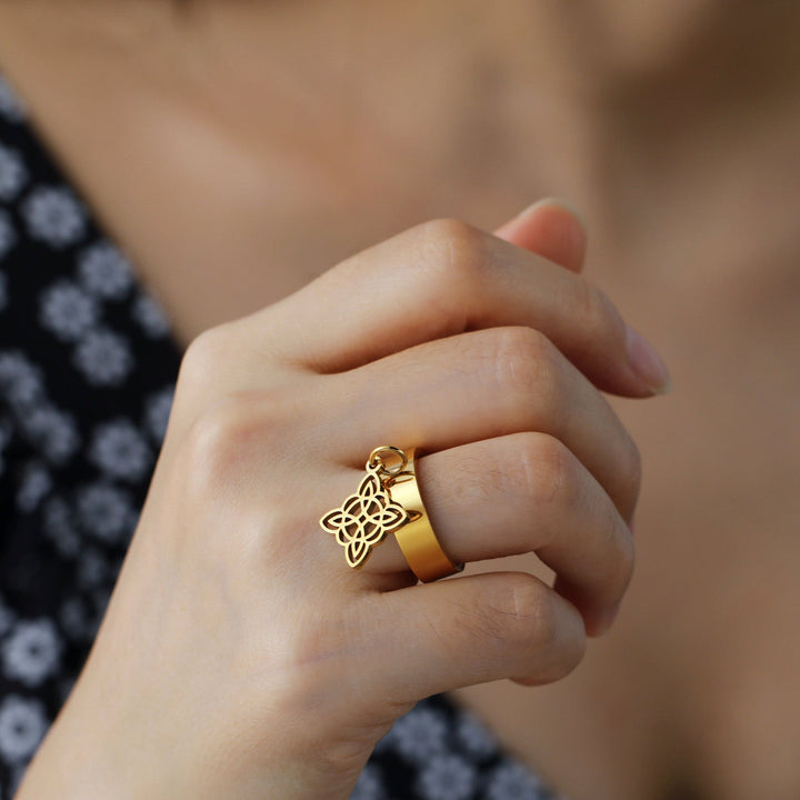 The photo shows a hand wearing THE GUARDIAN'S WITCHES KNOT DANGLE ring, made of stainless steel with Witch Knot element, in gold color