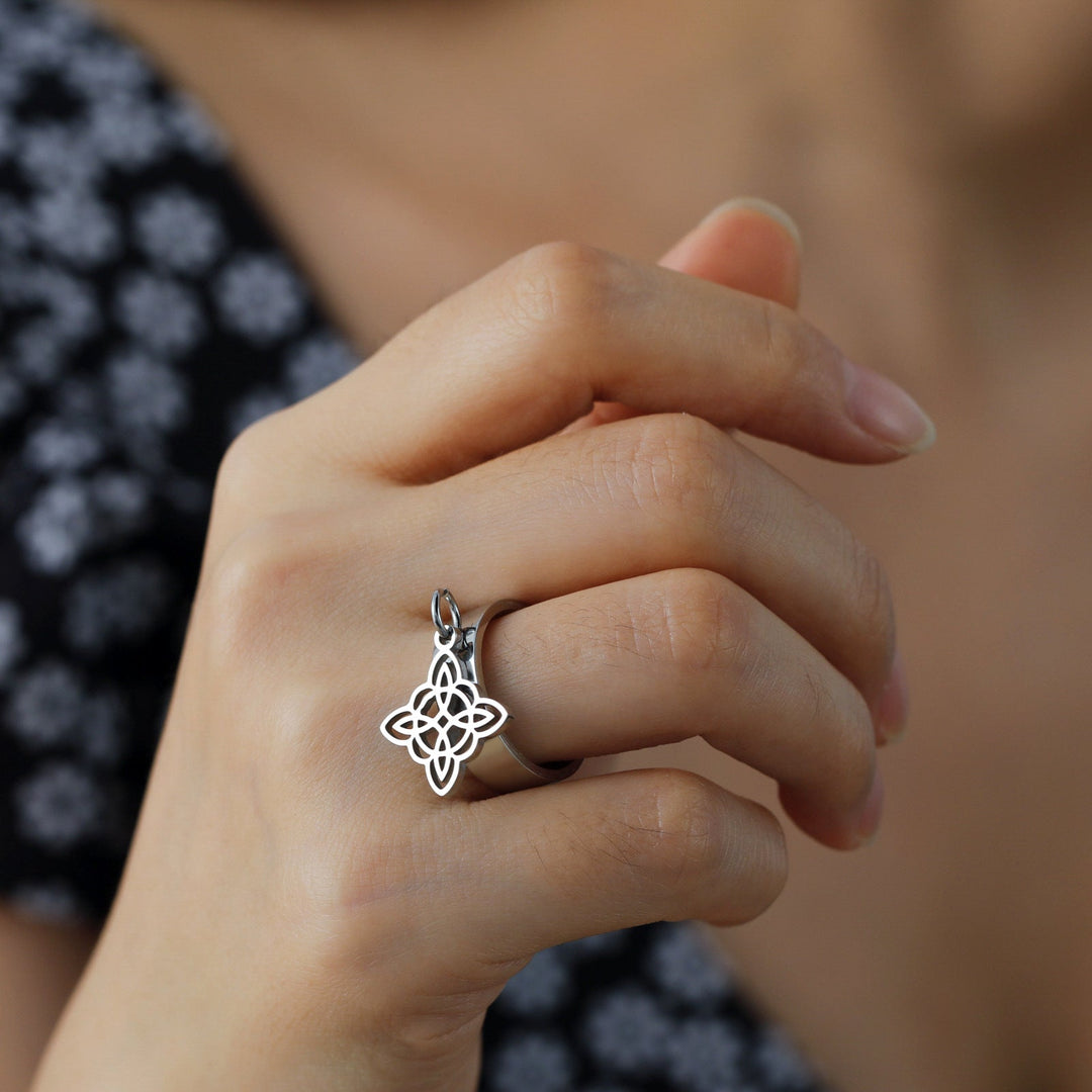 The photo shows a hand wearing THE GUARDIAN'S WITCHES KNOT DANGLE ring, made of stainless steel with Witch Knot element, in silver color