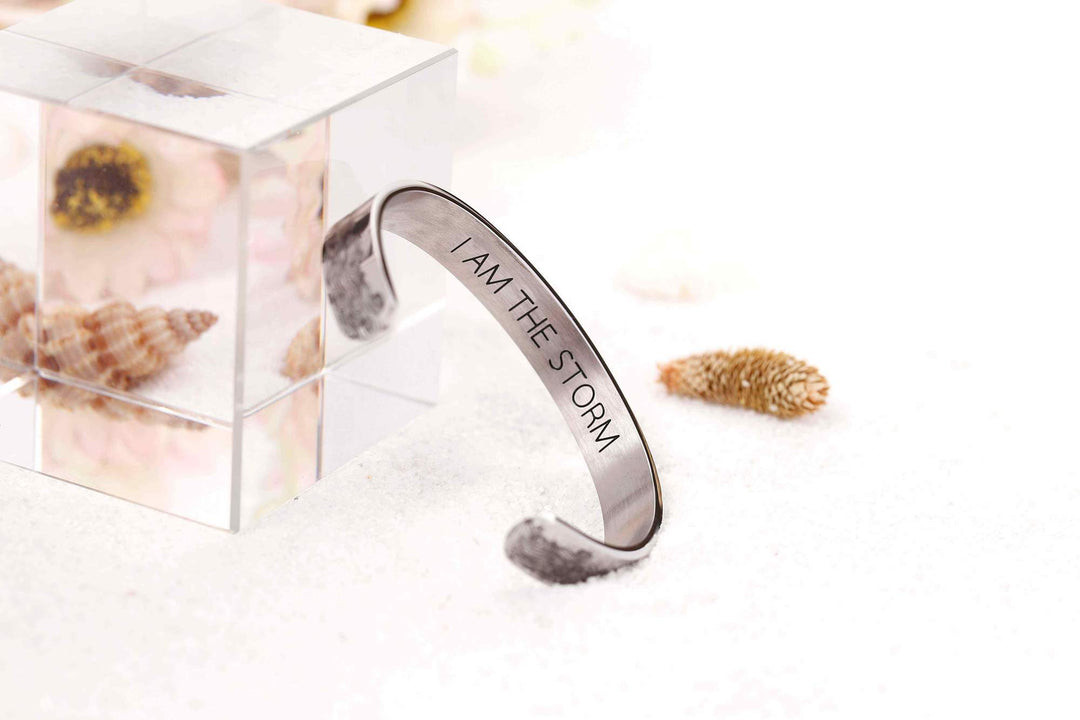 The image displays the "I AM THE STORM NEW" bracelet. The bracelet is made of titanium steel with 316L stainless silver plating, ensuring durability and resistance to fading, tarnishing, and corrosion. It features the inscription "I AM THE STORM NEW" on the inside. The bracelet has a diameter of 2.5 inches, a length of 6.2 inches, and a width of 3/8 inch, with adjustable openings to fit most wrists. The package includes a gift card, a gift bouquet, and a velvet bag for presentation.