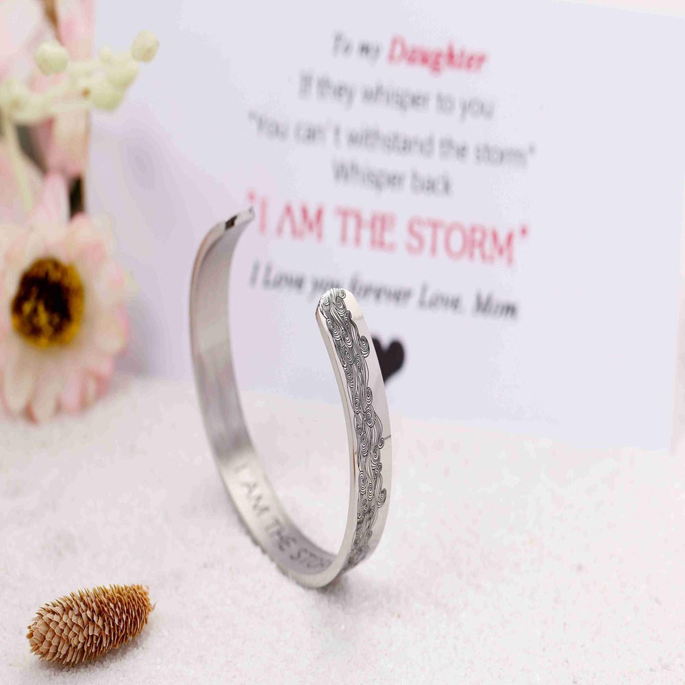 The image displays the "I AM THE STORM NEW" bracelet. The bracelet is made of titanium steel with 316L stainless silver plating, ensuring durability and resistance to fading, tarnishing, and corrosion. It features the inscription "I AM THE STORM NEW" on the inside. The bracelet has a diameter of 2.5 inches, a length of 6.2 inches, and a width of 3/8 inch, with adjustable openings to fit most wrists. The package includes a gift card, a gift bouquet, and a velvet bag for presentation.