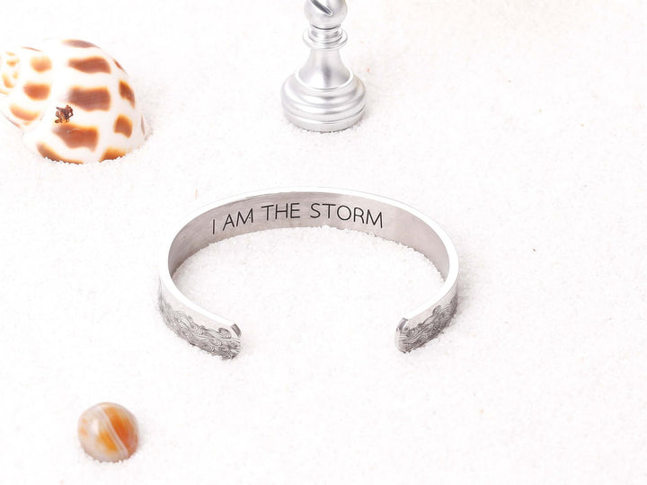 The image displays the "I AM THE STORM NEW" bracelet. The bracelet is made of titanium steel with 316L stainless silver plating, ensuring durability and resistance to fading, tarnishing, and corrosion. It features the inscription "I AM THE STORM NEW" on the inside. The bracelet has a diameter of 2.5 inches, a length of 6.2 inches, and a width of 3/8 inch, with adjustable openings to fit most wrists. The package includes a gift card, a gift bouquet, and a velvet bag for presentation.