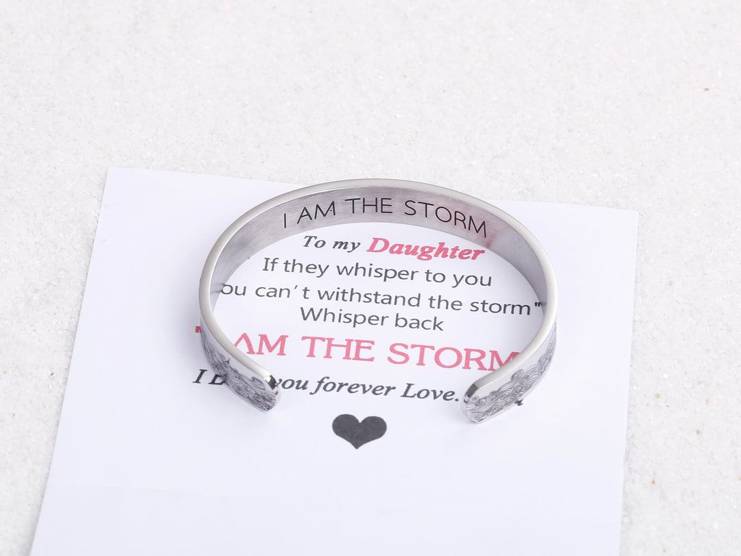 The image features the "I AM THE STORM NEW" bracelet, made of titanium steel with 316L stainless silver plating. The bracelet is durable and resistant to fading, tarnishing, corrosion, and allergic reactions. It has a diameter of 2.5 inches, a length of 6.2 inches, and a width of 3/8 inch, with adjustable openings to fit most wrists. The inside of the bracelet is inscribed with "I AM THE STORM NEW." Additionally, there is a gift card included in the package.